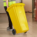 A person in a yellow vest pushing a Lavex yellow wheeled rectangular trash can.