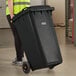 A woman in a safety vest pushing a Lavex black wheeled rectangular trash can.