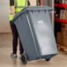 A woman in a yellow vest pushing a large grey Lavex rectangular trash can.