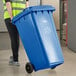 A woman in a yellow vest pushing a Lavex blue wheeled rectangular recycle bin.