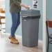 A woman standing next to a Lavex Gray square trash can with swing lid in a corporate office cafeteria.