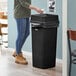 A woman standing next to a Lavex black square trash can with a white swing lid.