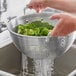 A person pouring broccoli into a Choice aluminum colander.