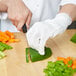 A person wearing Mercer Culinary white cut-resistant gloves cutting vegetables on a cutting board.