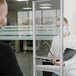 A man and a woman wearing masks stand in front of a MasterVision standing desk with a safety shield.