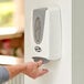 A person using a Noble Chemical white manual foam hand sanitizer dispenser on a wall.