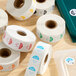 A collection of Noble 1-inch permanent day of the week clock label rolls in various colors on a wooden surface.