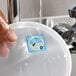 A dissolvable Monday day-of-the-week clock label being rinsed off a plastic container lid under running water.