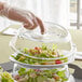 A hand wearing plastic gloves lifts a clear polycarbonate plate cover off a salad.