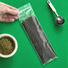 A hand using a Clear Line plastic bag to scoop spaghetti next to a bowl of herbs.