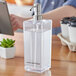 A person using an American Metalcraft clear plastic hand sanitizer dispenser on a table.