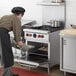 A man in a black chef's hat and apron using a Vulcan commercial gas range to put food into an oven.