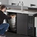 A man in a black shirt and jeans kneeling to open a silver keg in a black Beverage-Air beer cooler.