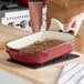 A man using a red Valor enameled cast iron roasting pan to cook a roast with herbs.