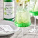 A glass of green liquid with a straw sitting on a table with a white bottle of Monin Premium Granny Smith Apple Fruit Syrup.