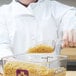 A person in a white coat using a Cambro clear polycarbonate scoop to pour macaroni into a container.