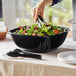 A person serving salad in a black Visions plastic bowl.