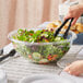 A person using tongs to serve salad from a Visions plastic bowl.