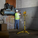 A man in a safety vest and hard hat using a Voltec LED work light on a yellow stand to check a pipe.