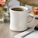 A white Acopa stoneware coffee mug with brown liquid and a spoon on a napkin.