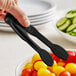 A person holding Thunder Group black flat grip tongs over a bowl of tomatoes.