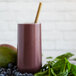 A red cylindrical Pitaya Foods Organic Acai and Guarana smoothie pack on a table next to a glass of purple smoothie with a straw.