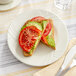 An Acopa ivory stoneware plate with a sandwich, avocado, and tomato slices on a table.