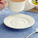 A hand holding a cup of coffee over an ivory stoneware saucer on a table with a bowl of fruit.