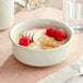 An Acopa ivory stoneware bowl filled with oatmeal and raspberries on a white surface with a black border.