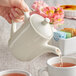 A person pouring water into a Tuxton eggshell white teapot.