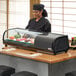 A woman in a chef's hat standing behind a counter with an Emperor's Select Countertop Refrigerated Sushi Display Case full of sushi.