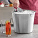 A person pouring ice into a Vollrath stainless steel utility bucket full of ice.