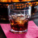 A Carlisle clear plastic tumbler with ice and brown liquid on a napkin.
