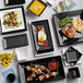 A table with Acopa matte black square stoneware plates and a black tray of food.