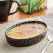 A black Acopa oval stoneware dish filled with creme brulee on a table.