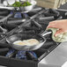 A hand using a green cloth to hold a Vollrath Wear-Ever non-stick fry pan with eggs cooking on a stove.