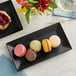 A rectangular matte black Acopa stoneware platter with macarons on a table.