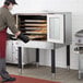 A man wearing a mask and gloves using a metal pan to put food in a Cooking Performance Group deep depth convection oven.