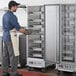 A man wearing a striped apron and gloves opening a Main Street Equipment heated holding cabinet to reveal trays of food.