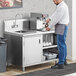 A man in a white shirt and blue jeans standing at a Regency stainless steel beverage table in a professional kitchen.