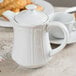 A white Tuxton Chicago teapot with a lid on a table with a cup.