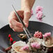 A hand holding Tablecraft culinary tweezers over a plate of food.