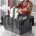 A man in a brown apron putting a silver pot into a Lavex black industrial tote with attached lid.