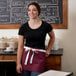 A woman wearing a burgundy waist apron with natural webbing and 3 pockets standing in front of a chalkboard.