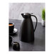 An Alfi black plastic vacuum insulated carafe on a table with a cup of white liquid.