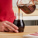 A person pouring brown syrup into a Vollrath teardrop syrup server jar.