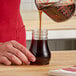 A person pouring brown syrup from a measuring cup into a clear teardrop-shaped jar.
