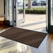 A brown Lavex Needle Rib entrance mat in front of a building's glass doors.