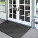 A black Lavex Chevron Rib entrance mat with a charcoal border on a white floor in front of a double glass door.