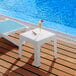 A Lancaster Table & Seating white side table on an outdoor patio next to a pool with a drink on it.
